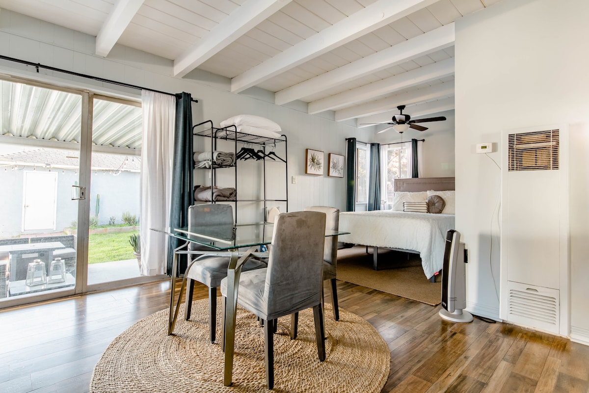 The studio features a spacious layout with wooden flooring, a queen bed, and a glass dining table accompanied by two chairs. A sliding glass door provides access to the outdoor area. Shelving units are neatly arranged, and a ceiling fan is present above for comfort.