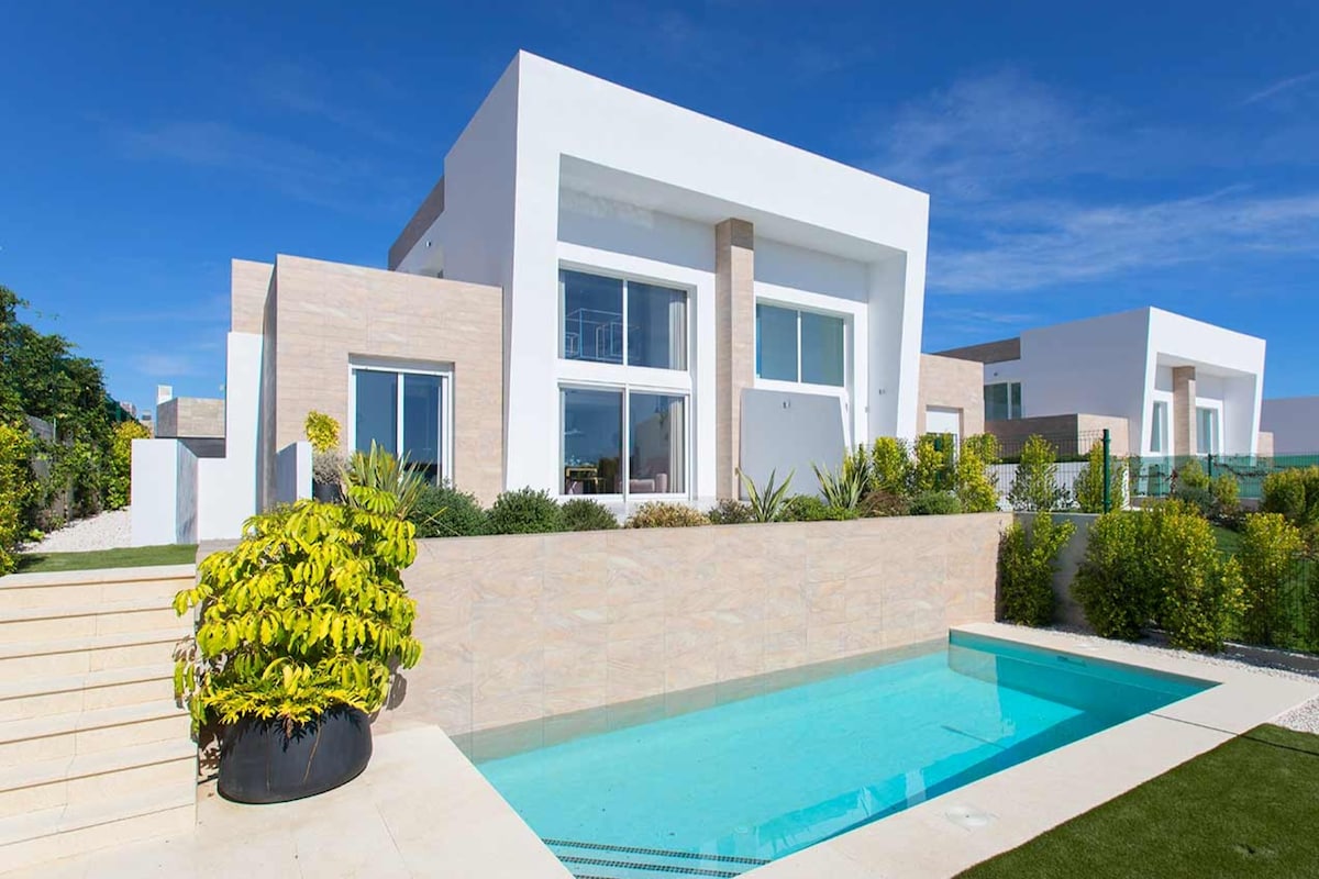 A modern villa showcases clean lines and large glass windows, surrounded by landscaped gardens. A private pool reflects the blue sky, while steps lead down to the entry, enhancing the inviting outdoor space.