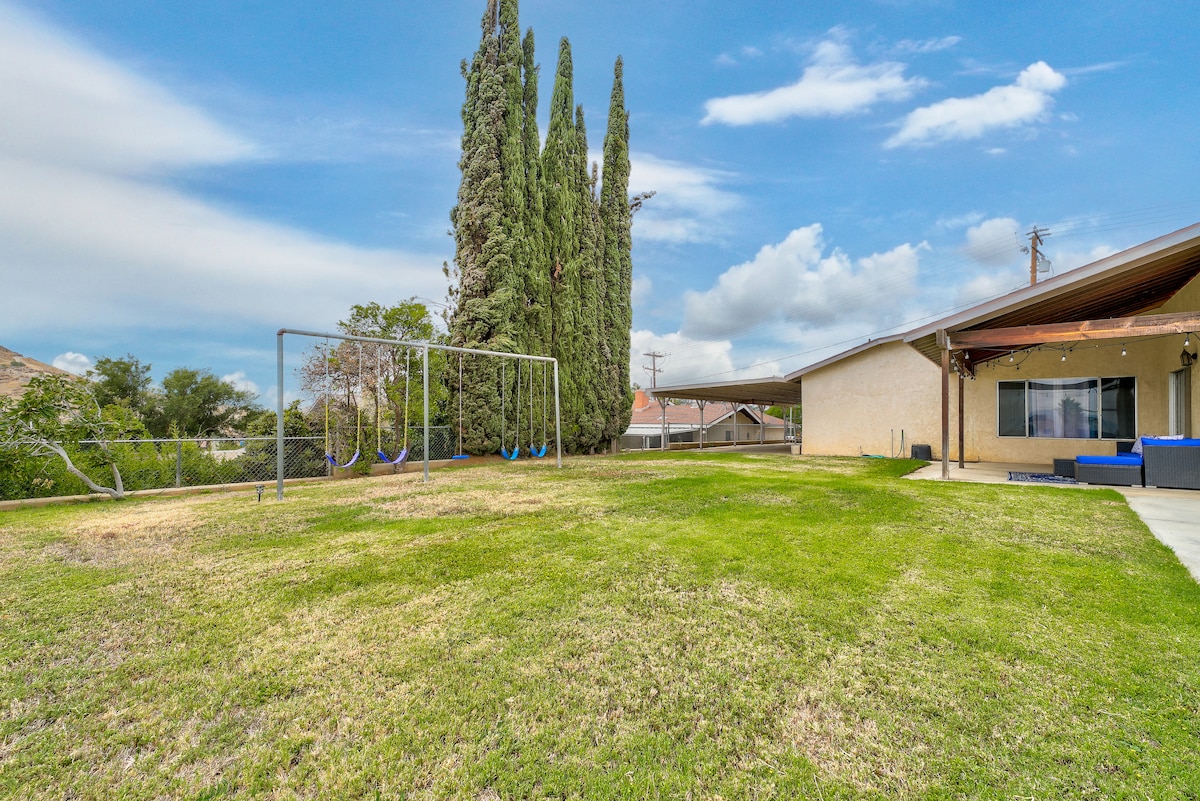 A spacious backyard is shown, featuring a green lawn and a children's swing set. Tall trees provide a natural border, with the home's patio area visible in the background. The landscape is open and well-maintained, inviting outdoor activities.