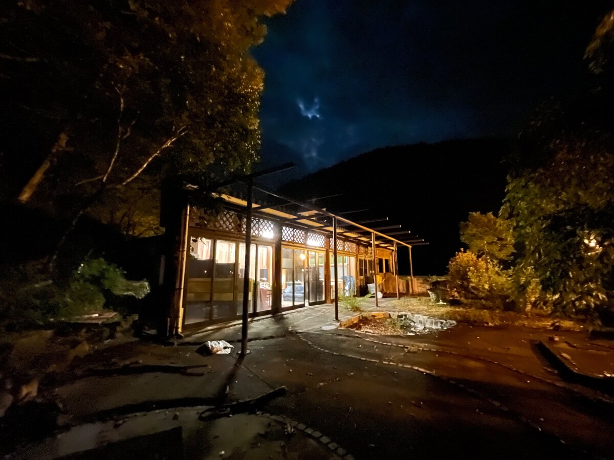 The exterior of a wooden building is illuminated against a dark sky, showcasing large glass windows and an expansive porch. Surrounding trees and plants create a natural setting, hinting at the peaceful atmosphere of the area at night.