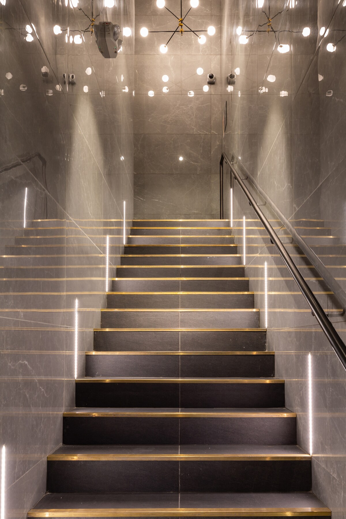 A modern staircase is lit by recessed lighting along each step, with chrome handrails providing support. The walls are clad in reflective tiles, enhancing the overall brightness, while a series of pendant lights hangs from the ceiling above.