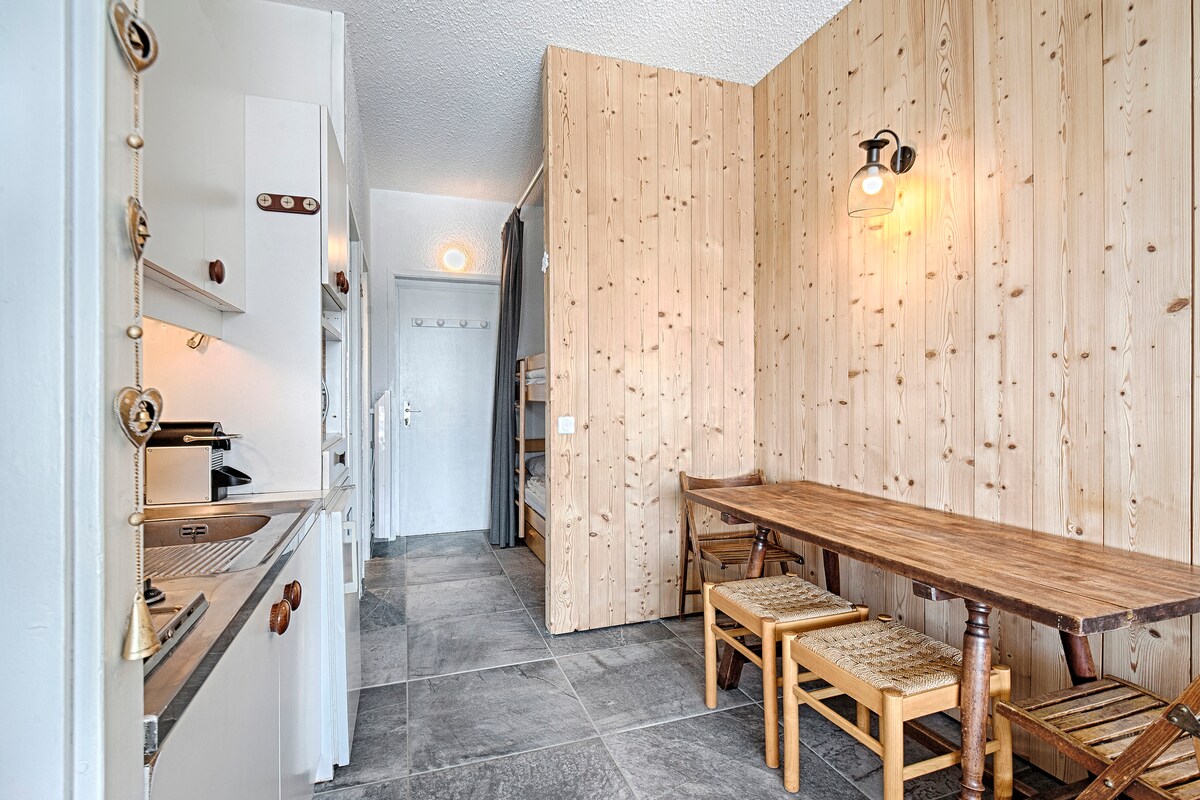 The kitchenette is equipped with modern appliances, including an induction cooktop and refrigerator. A rustic wooden dining table with two woven chairs is positioned adjacent to the kitchen area, surrounded by warm wood paneling. The space is brightened by a light fixture on the wall.