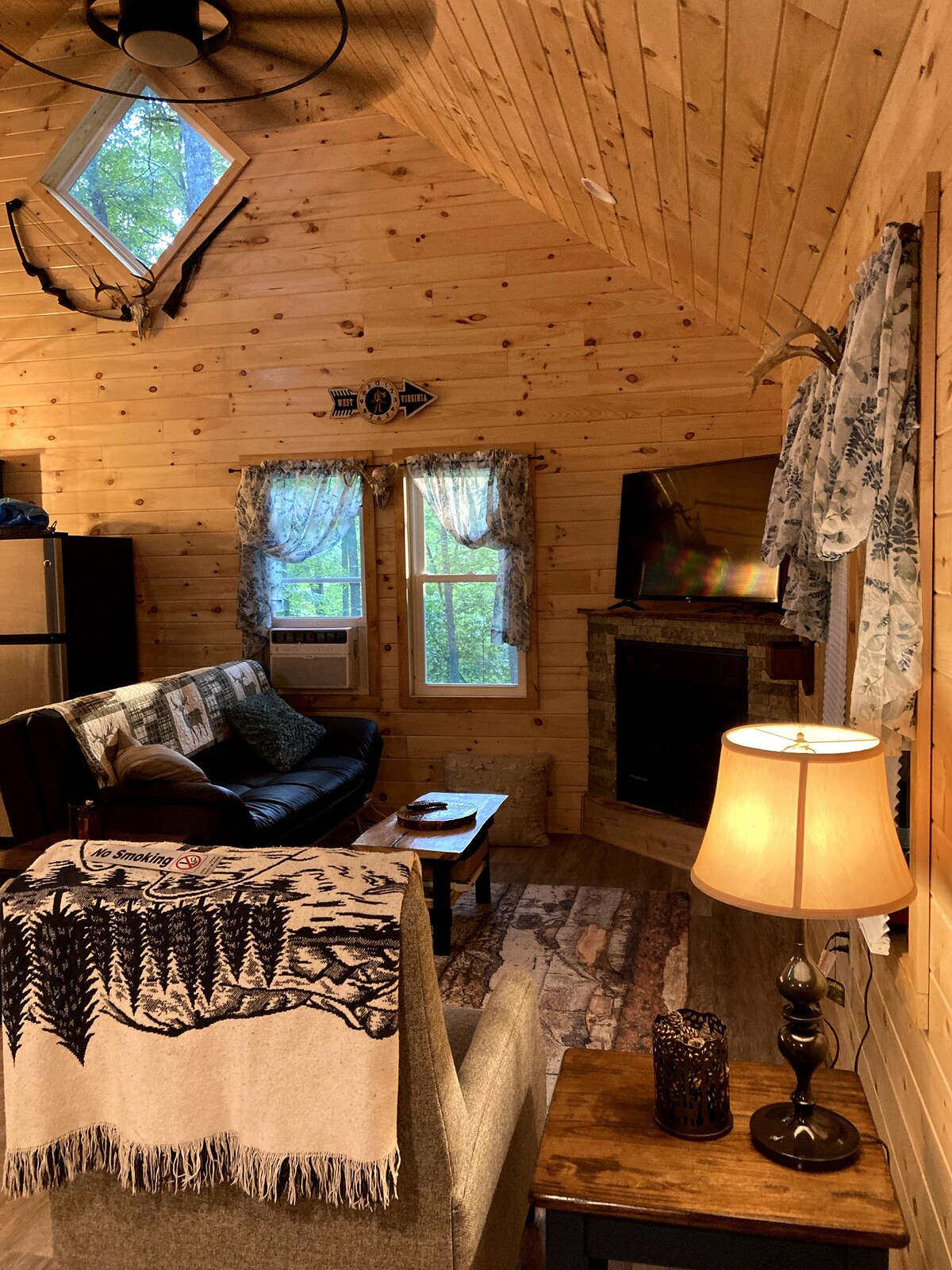 A cozy living area features wooden walls and a sloped ceiling. A dark leather sofa is adorned with a decorative throw, while a lamp provides soft illumination. Two windows with lace curtains allow natural light to enter, complementing the rustic decor and warmth of the space.
