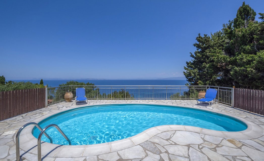 A kidney-shaped private pool is outlined by a stone terrace, featuring two blue sun loungers. The pool offers expansive views of the Ionian Sea under a clear blue sky, surrounded by greenery and a wooden fence, providing a serene and inviting outdoor space.