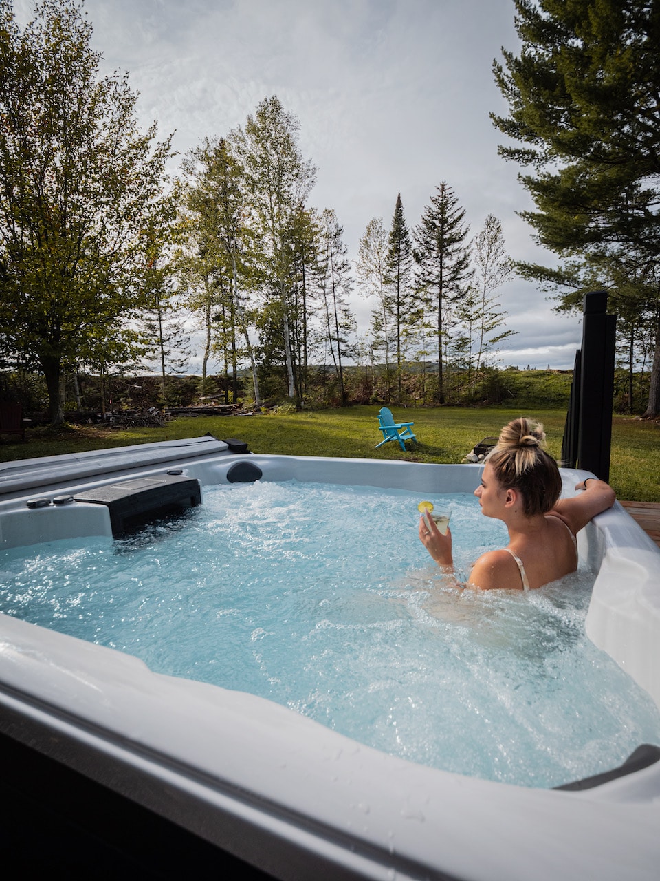 A hot tub is set on a wooden deck, with a person relaxing inside. Surrounding trees provide a natural backdrop, and a clear blue sky is visible above. Comfortable outdoor seating is seen nearby, enhancing the tranquil outdoor environment.
