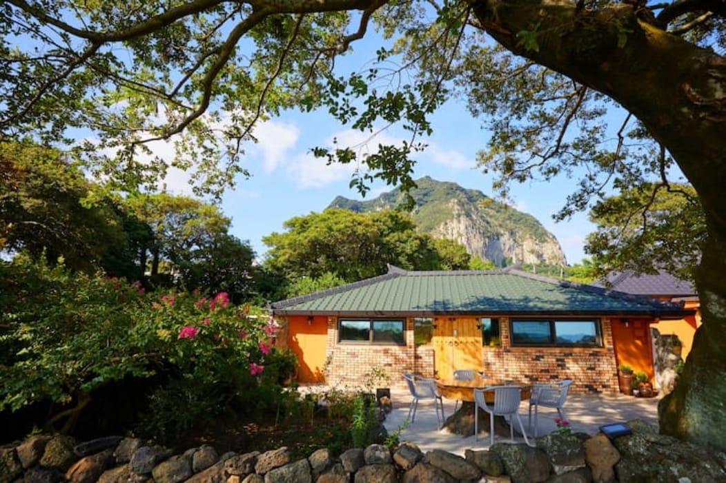 A charming house is surrounded by lush greenery, with a view of a prominent mountain in the background. The exterior features a mix of brick and vibrant orange walls, complemented by outdoor seating arranged on a stone patio under the shade of large trees.
