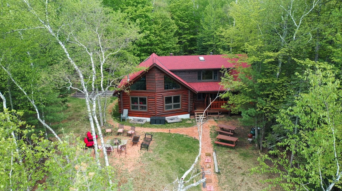 A full log home is nestled among lush greenery, surrounded by tall trees. The red roof contrasts with the natural tones of the wood. Outdoor seating, including picnic tables and a fire pit area, is accessible, inviting relaxation in a peaceful, secluded setting.