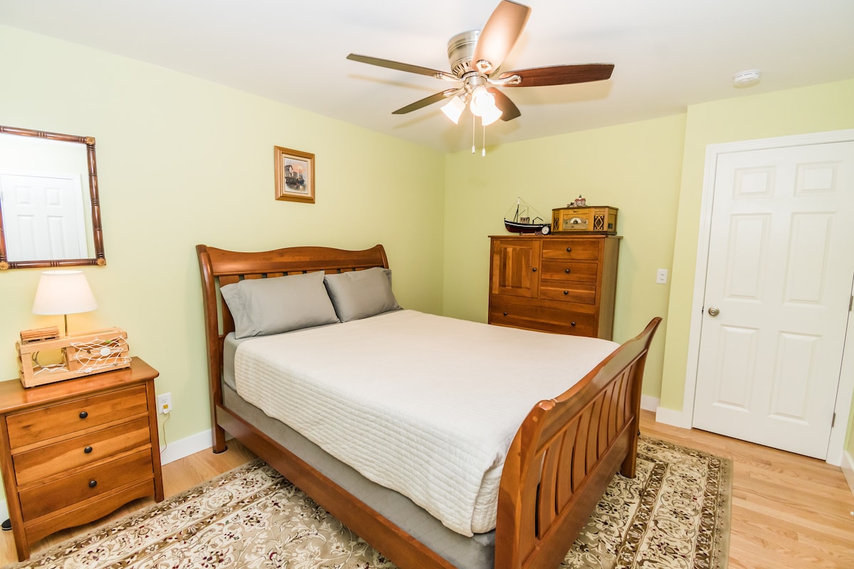 A private bedroom features a wooden bed with a light-colored bedspread and two pillows. A ceiling fan is present above, and a mirror reflects the warm tones of the room. A wooden dresser and a small bedside table provide storage, with natural light illuminating the space.