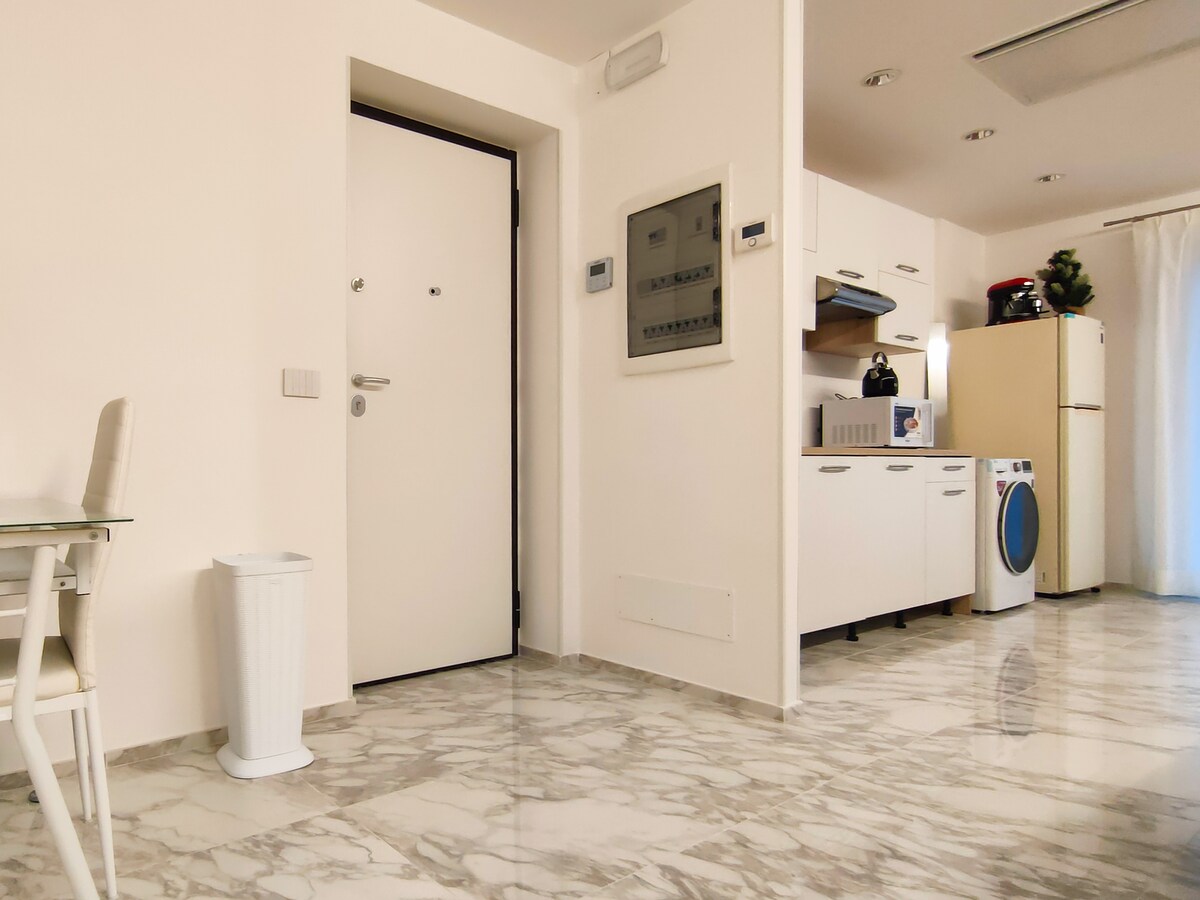 An open area presents a clean and modern kitchen, equipped with essential appliances. The entrance door is visible alongside a compact dining table with chairs. Light-colored marble flooring reflects the bright, neutral tones of the space.