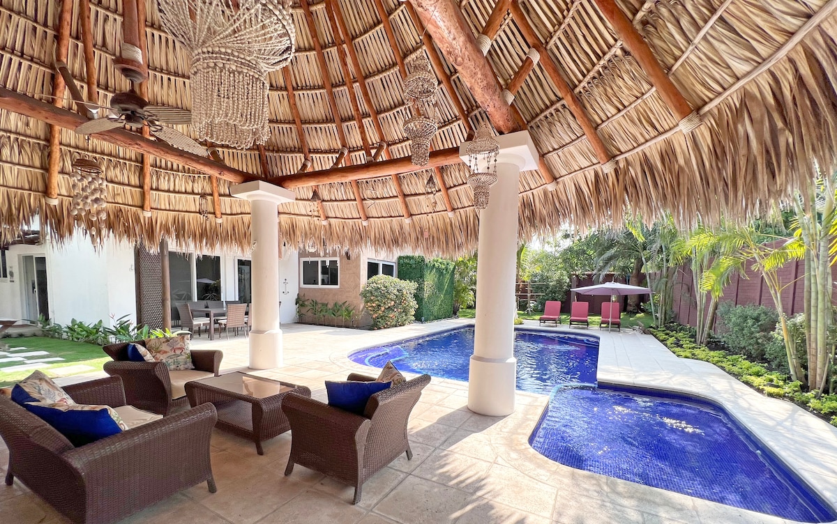 A spacious patio features a traditional palm-thatched roof, providing shade over comfortable seating. A glistening pool is visible, surrounded by lush greenery. Nearby, lounge chairs and an outdoor dining area enhance the relaxing atmosphere, inviting guests to enjoy the tropical surroundings.