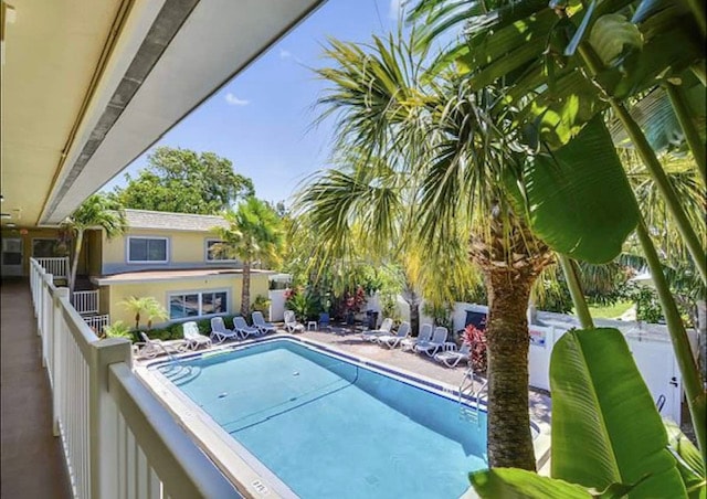 A view from the second-floor balcony overlooks a swimming pool surrounded by lush palm trees and tropical plants. Several lounge chairs are positioned around the pool area, inviting relaxation in a sunny atmosphere.