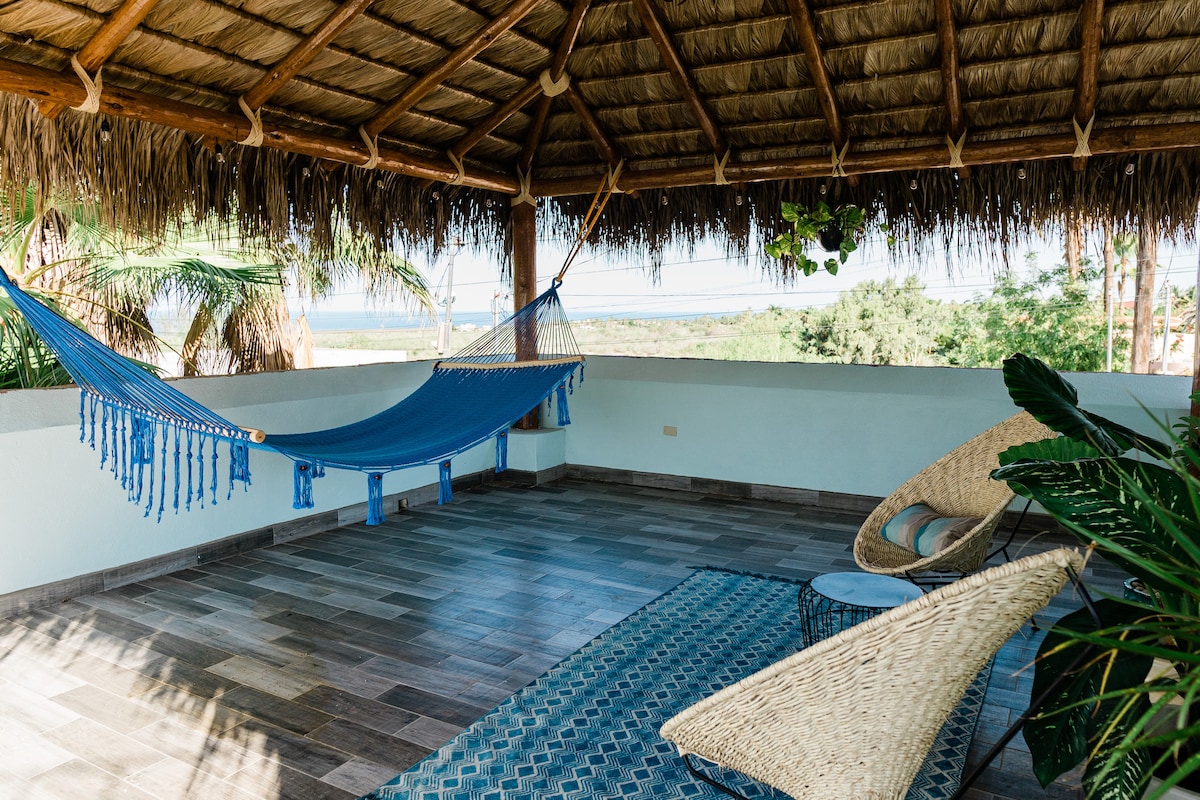 A palapa-covered terrace features a blue hammock and two woven seating chairs. A patterned rug lies beneath, while potted plants add greenery to the space. The serene ocean and mountain views are visible in the background, creating a calming outdoor atmosphere.