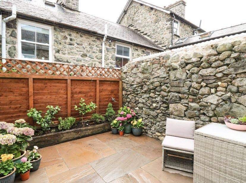An enclosed courtyard features a stone wall providing privacy. Potted plants and flowers are arranged along the edges, while a small cushioned chair and a woven table offer a cozy seating area amidst the greenery. The paved floor adds a natural touch.