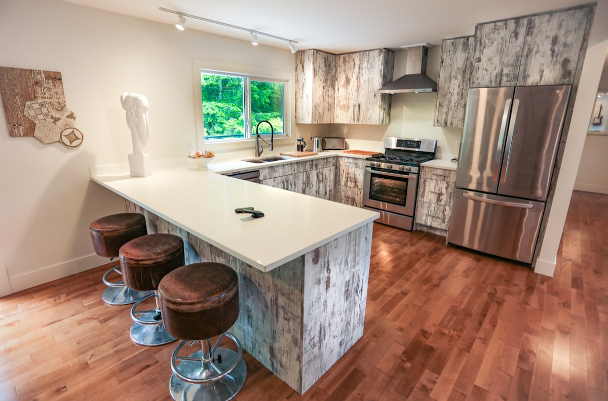A modern kitchen is displayed featuring gray-toned cabinetry with a wood-like finish and stainless steel appliances. A spacious countertop bar includes three round stools with brown leather seats. Natural light enters through a nearby window, illuminating the space.