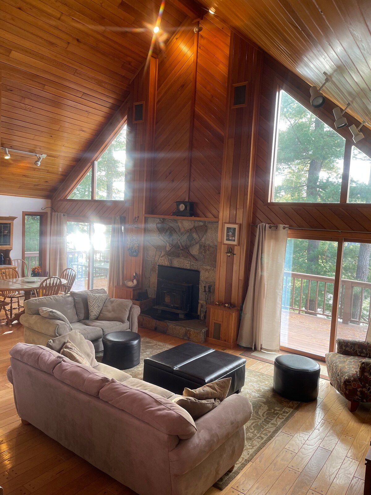 A spacious living area features high cathedral ceilings adorned with wooden beams. A stone fireplace serves as a focal point, complemented by large windows that offer views of the surrounding trees. Comfortable seating is arranged invitingly around a central coffee table.