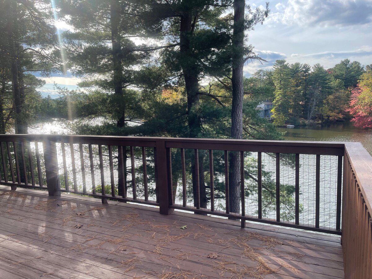 A wooden deck overlooks the tranquil waters of Lake Garda, surrounded by tall pine trees. Soft sunlight filters through the branches, casting gentle shadows on the deck surface. The scenery reflects a calm, natural setting ideal for relaxation.