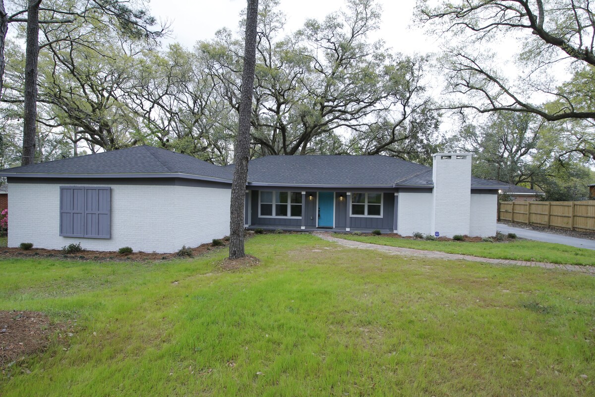 The exterior of the ranch-style home showcases a modern façade with light-colored brick and a dark roof. Large windows invite natural light, while a welcoming blue front door offers access to the interior. The surrounding yard features green grass and mature trees.