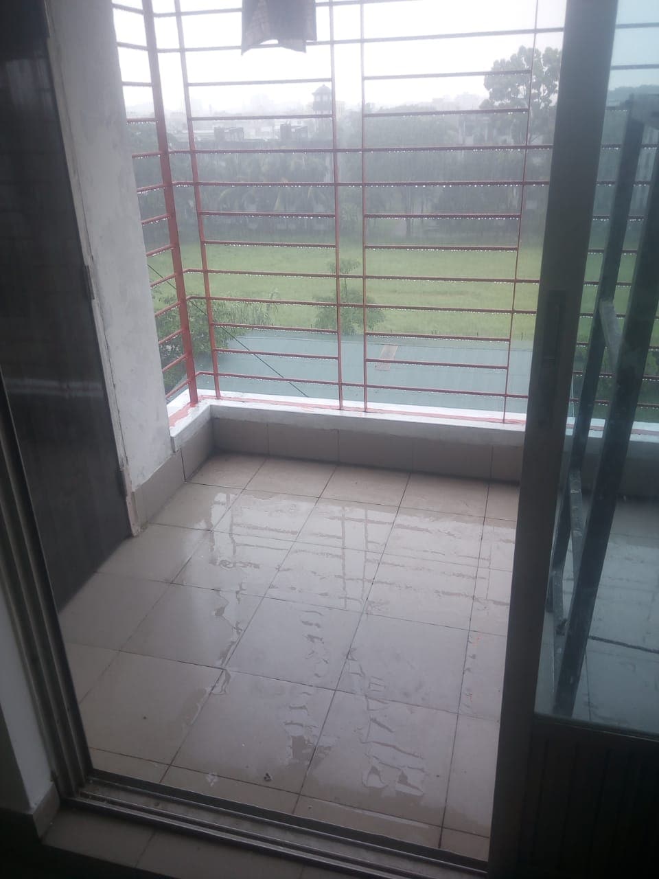 A small balcony area is visible with tiled flooring reflecting dampness from recent rain. A red metal railing borders the space, revealing a view of a green landscape beyond. Glass sliding doors lead into the interior, enhancing the connection between indoor and outdoor.