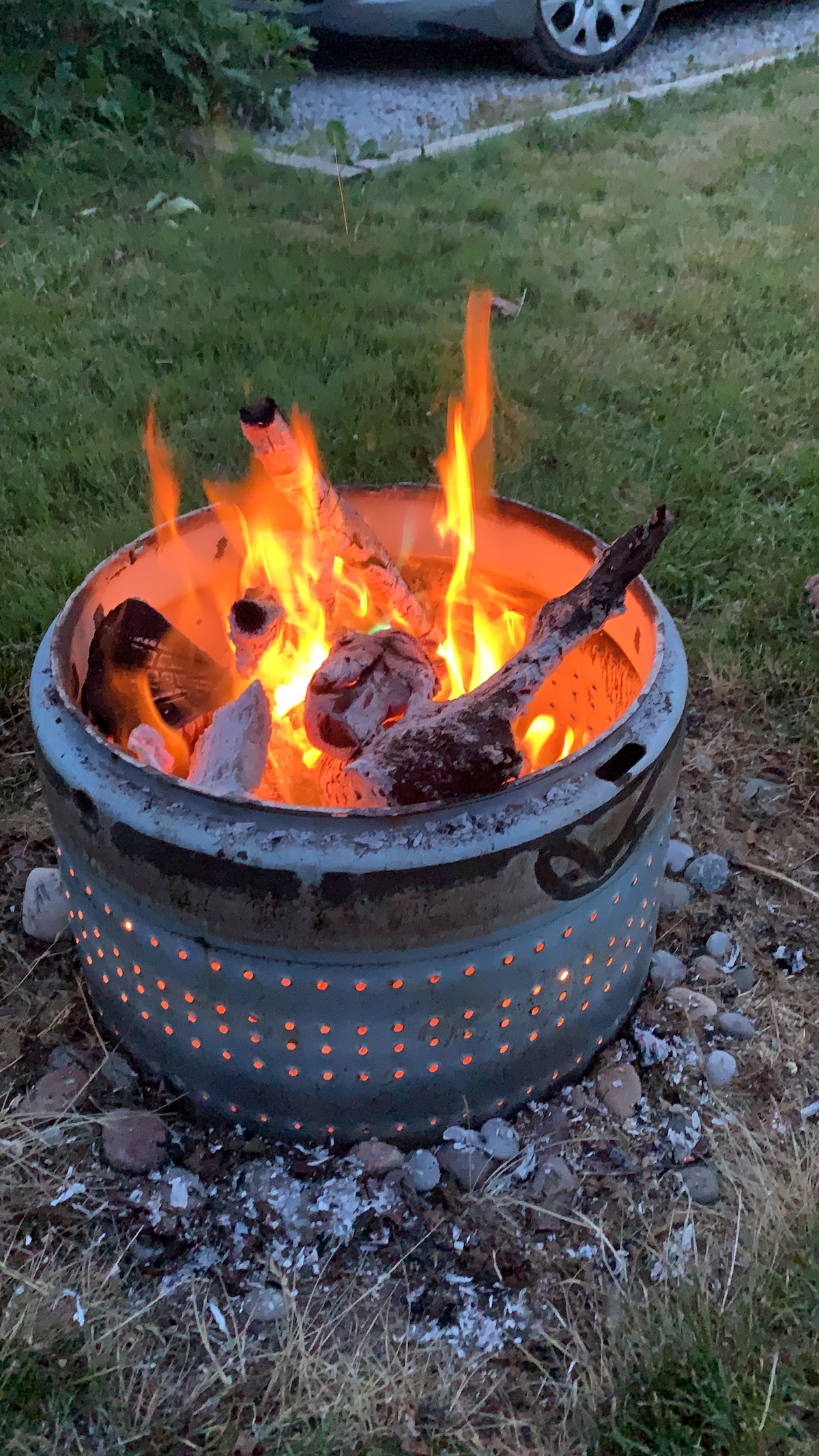 A campfire is lit within a metal fire ring, surrounded by grass and small stones. Flames dance above charred wood, providing warmth and ambiance to the outdoor setting. A vehicle is visible in the background, indicating proximity to access points.