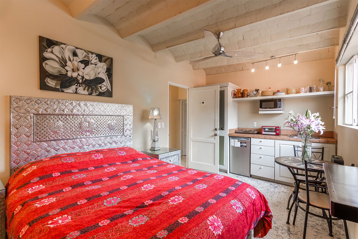 A spacious sleeping area features a large bed with a vibrant red bedspread, complemented by decorative wall art. A kitchenette is visible in the background, equipped with modern appliances. Natural light flows in through a window, enhancing the warm ambiance of the room.