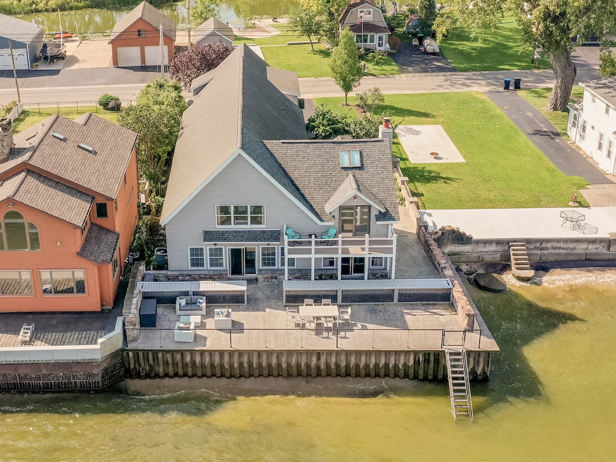 Aerial view of a lakefront house, showcasing a spacious deck with outdoor furniture, surrounded by lush greenery and nearby homes. The water's edge features a sandy bottom, and a staircase leads down to the shoreline, offering direct access to Lake Ontario.