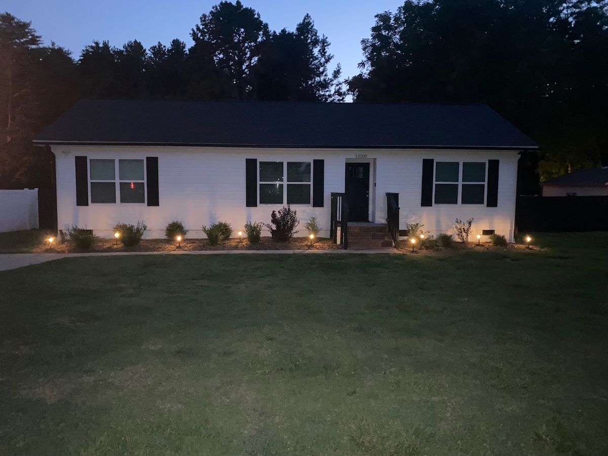The front of the house is illuminated in the evening, showcasing a single-story structure with white siding and dark green shutters. Landscape lighting highlights the garden beds along the pathway, and a well-kept lawn extends to the front steps.