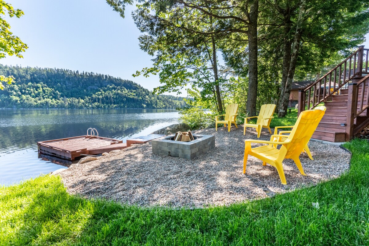 A lakeside area is featured with a fire pit surrounded by four yellow chairs on a smooth gravel surface. The private dock extends into the shimmering water, and lush greenery lines the shore, providing a serene backdrop.