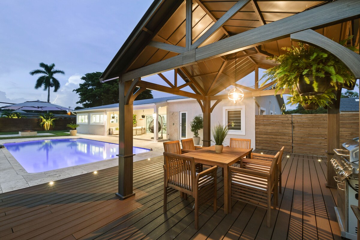 An outdoor seating area is positioned under a sheltered patio, featuring a wooden dining table surrounded by matching chairs. The space overlooks a heated pool, with palm trees and lush greenery in the background, creating a relaxing ambiance.