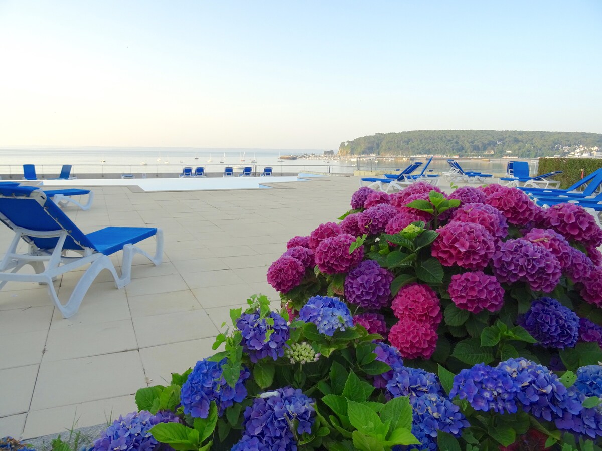Vibrant hydrangeas in pink and blue blossom in the foreground, while a swimming pool is visible beyond. Sun loungers are arranged neatly on the patio, offering a view of the calm bay and distant shoreline under a clear sky.