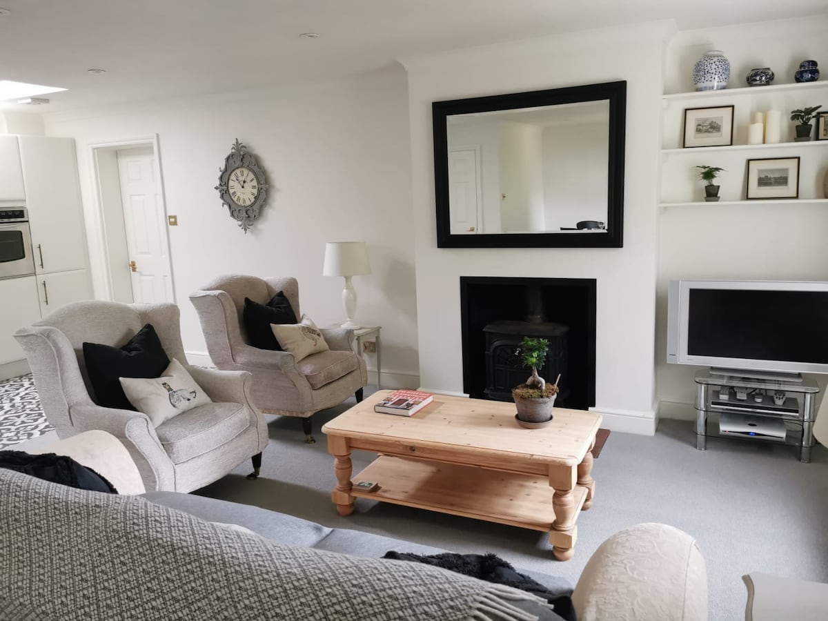 The living area features a spacious layout with a comfortable large sofa and two armchairs. A wooden coffee table holds a small potted plant, while a large wall mirror reflects natural light. A wall-mounted TV is positioned beneath a simple fireplace, contributing to a cozy atmosphere.