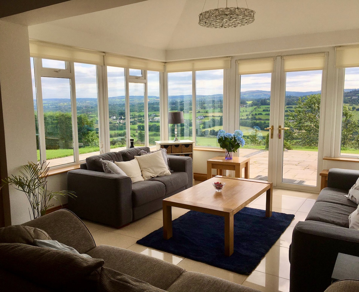 A bright and airy living space is filled with natural light, featuring large glass windows that offer sweeping views of the surrounding countryside. Comfortable sofas are arranged around a wooden coffee table, and a stylish light fixture hangs overhead, enhancing the inviting atmosphere.