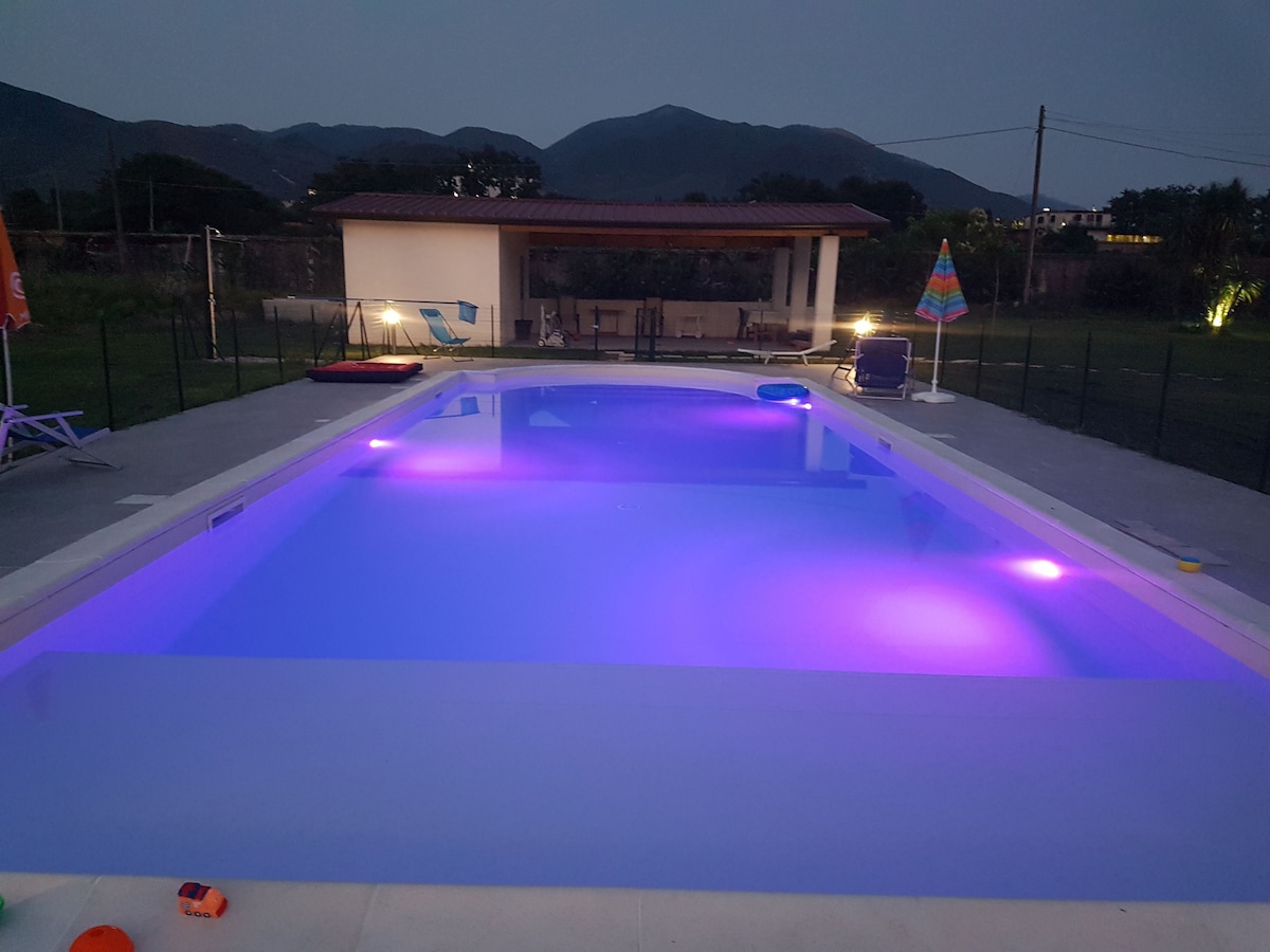 A large swimming pool is illuminated with purple lighting, creating an inviting atmosphere during twilight. Surrounding the pool, lounge chairs are arranged near a shaded area, with distant mountains providing a scenic backdrop. The overall setting is tranquil and spacious.