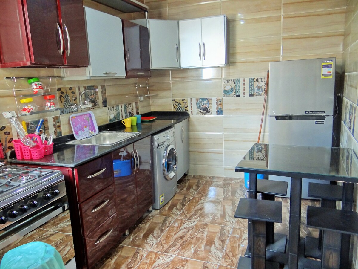 The kitchen features modern cabinetry in dark tones, complemented by a silver refrigerator. A stainless steel sink is positioned beside a washing machine. A small dining table with high stools occupies one corner, while decorative tiles add visual interest to the walls.
