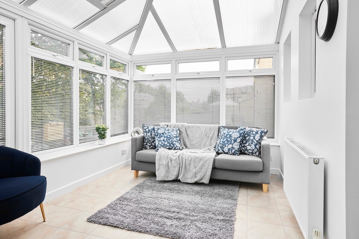 A bright sunroom features large windows adorned with blinds, providing ample natural light. A comfortable gray sofa is draped with a soft blanket and accented with blue-patterned cushions. A plush area rug sits beneath the sofa, while a small plant adds a touch of greenery.