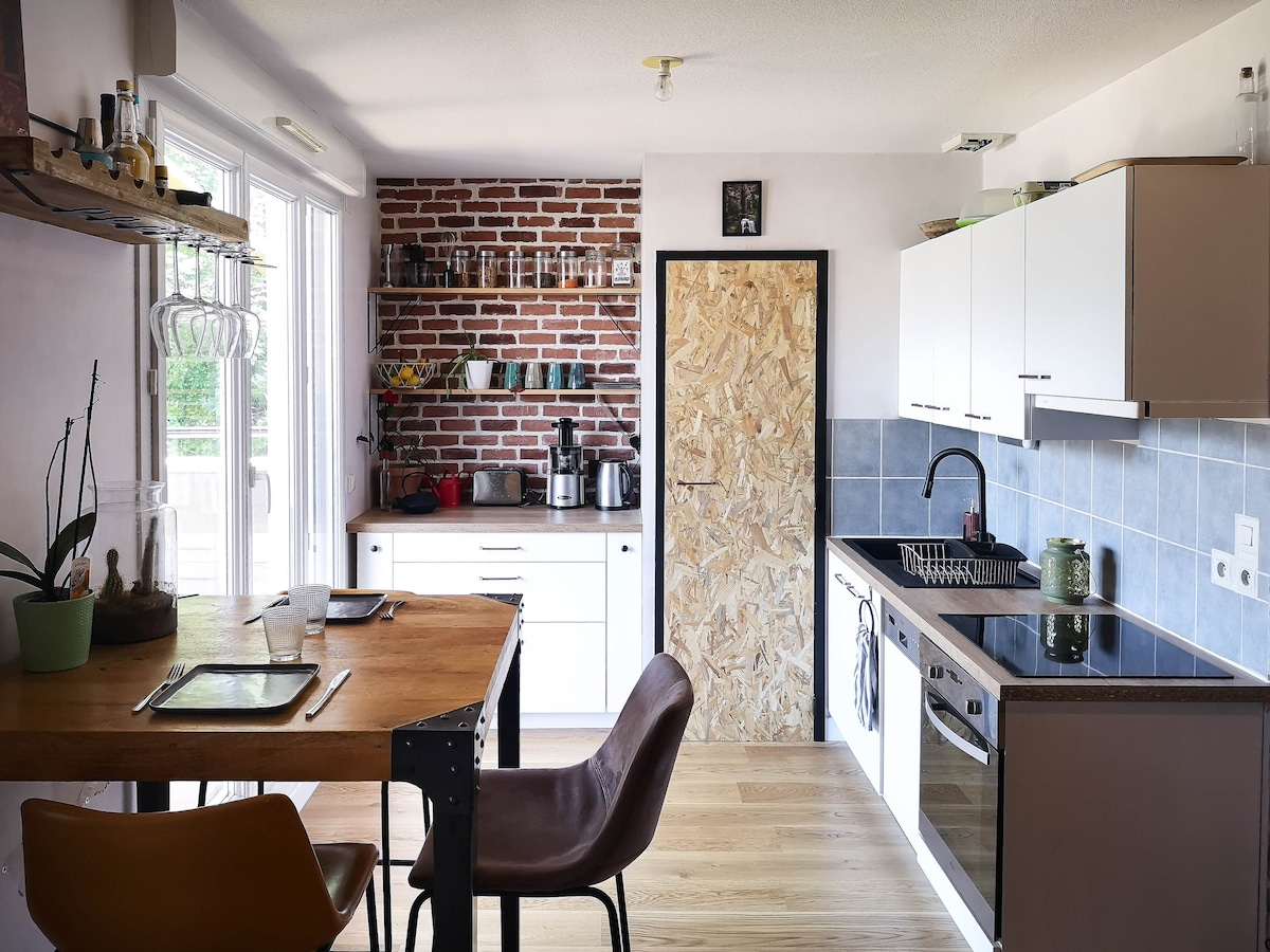 A modern kitchen features a combination of white cabinetry and exposed brick accents. An island with seating is situated beside a window, allowing natural light to illuminate the space. Equipped with an oven, stovetop, refrigerator, and coffee maker, the kitchen promotes a functional cooking environment.