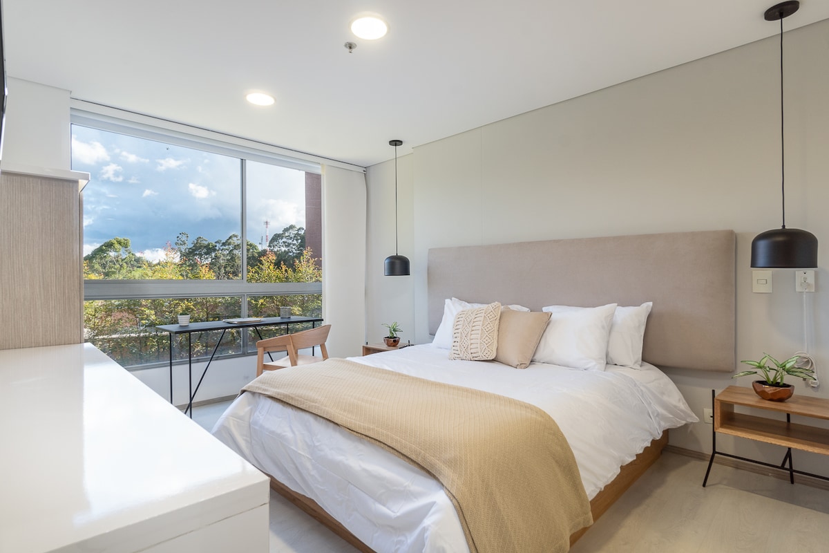 A bright bedroom features a spacious bed dressed in white linens and a light beige throw. Natural light enters through large windows, with greenery visible outside. A small desk and chair are positioned next to the bed, complementing the neutral color scheme of the room.