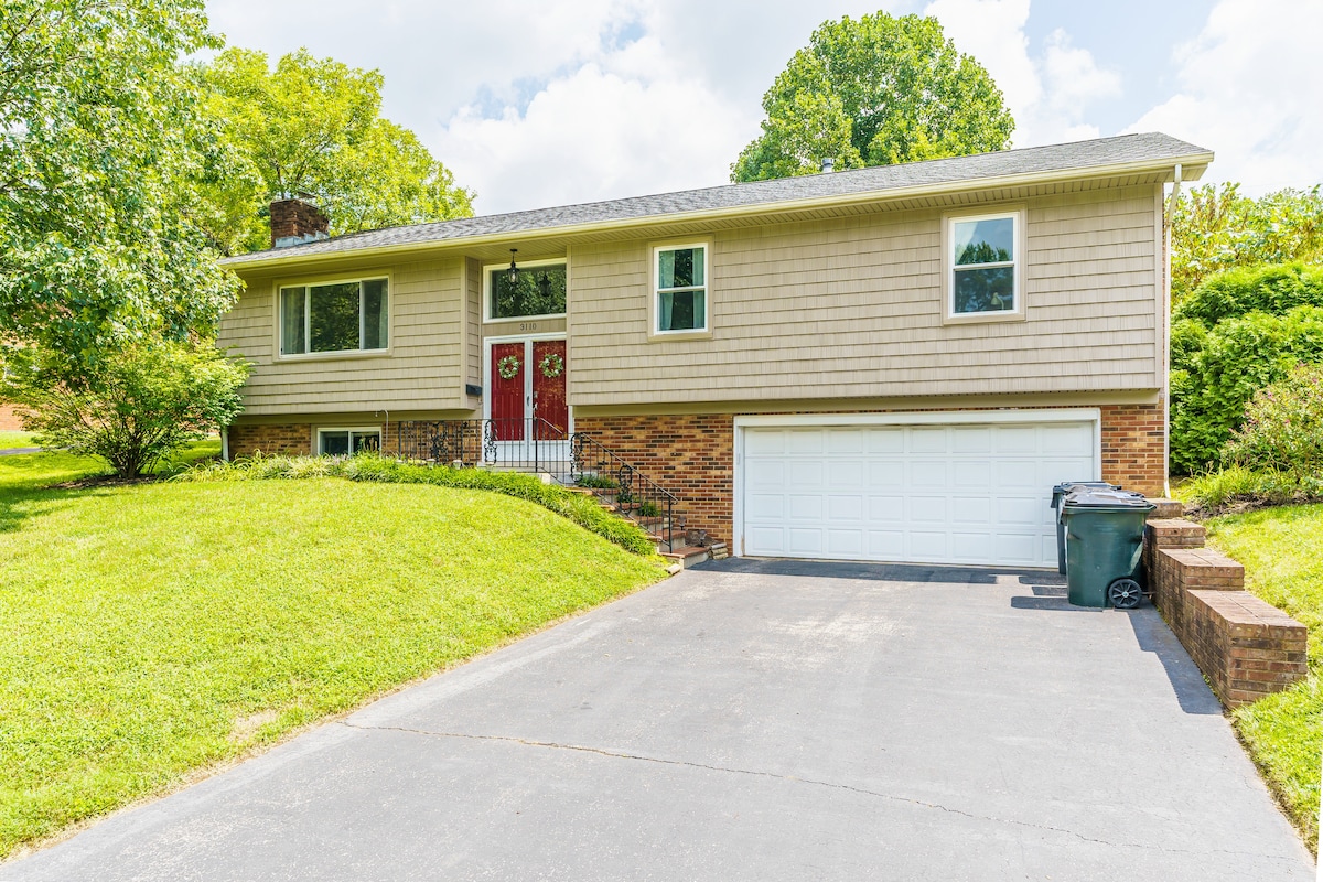 A split-level home is set against a vibrant green landscape, featuring a manicured lawn and a concrete driveway leading to a garage. The exterior showcases light-colored siding and brick accents, with inviting double doors framed by decorative wreaths.