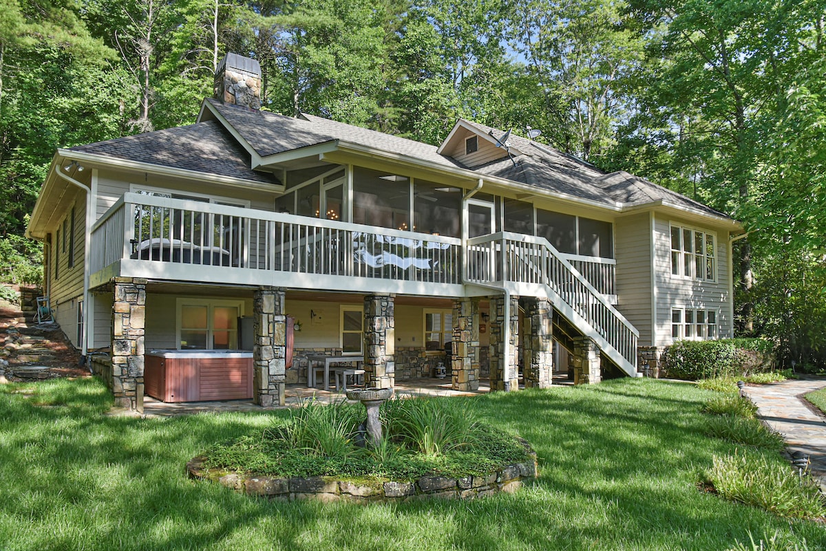 The exterior of the lakefront home features a large screened-in porch accessible by a staircase, surrounded by lush green grass. Native stonework supports the structure, enhancing its rustic appeal. A private hot tub is situated on the lower level, inviting relaxation.