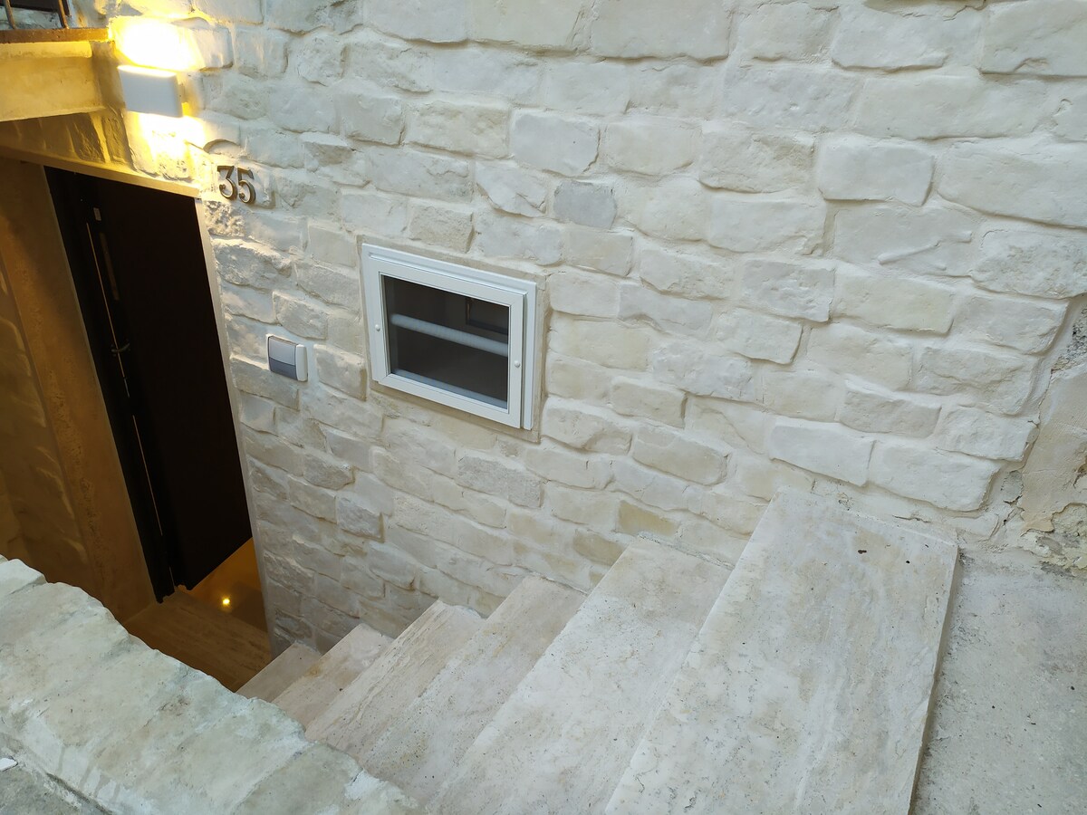 A stone staircase leads down to a doorway, accompanied by a small window embedded in the textured light-colored wall. Soft lighting highlights the steps, creating a welcoming entrance to the tranquil space below.