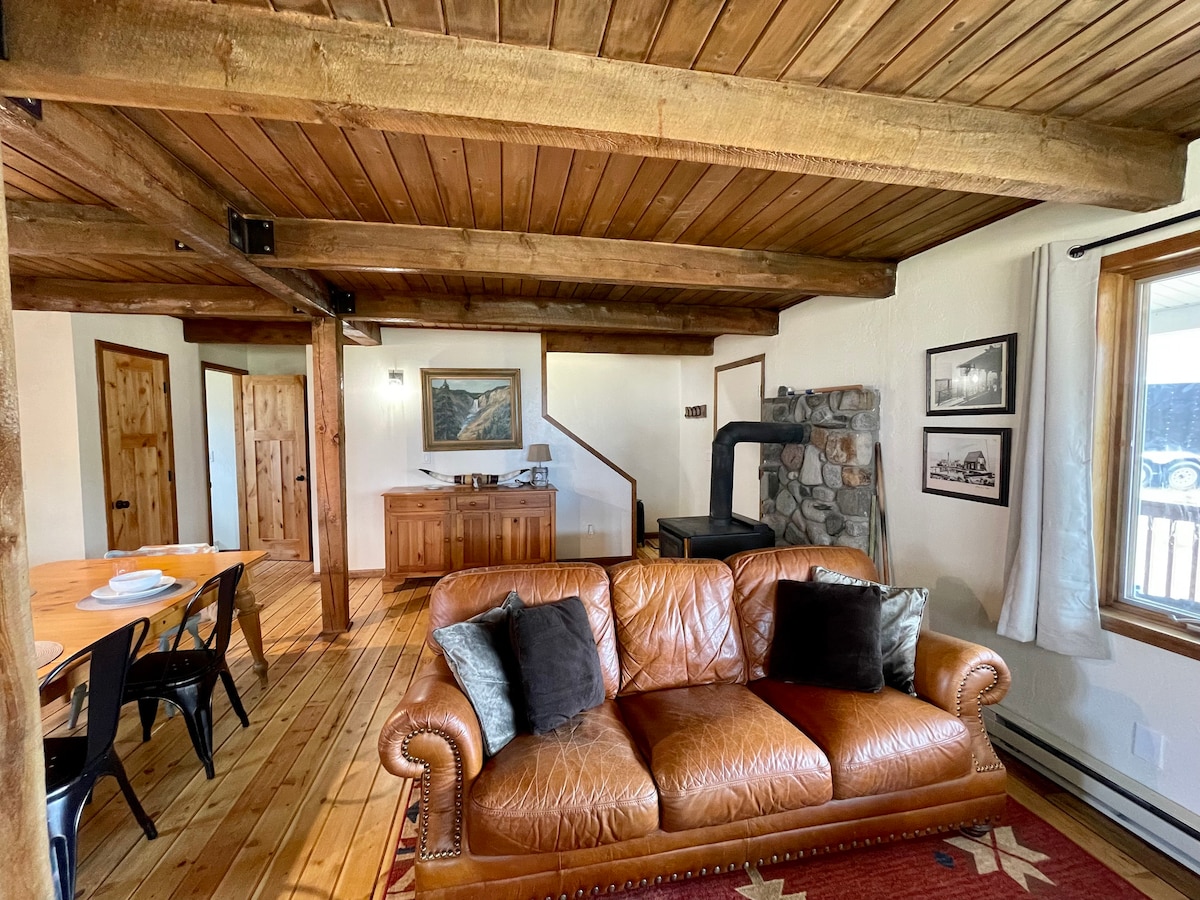 A spacious living area features a leather couch with decorative pillows, set on a warm wooden floor. Exposed beams adorn the ceiling, while a rustic wood stove and a wooden sideboard add character. Nearby, a dining table offers a place for meals and gatherings.