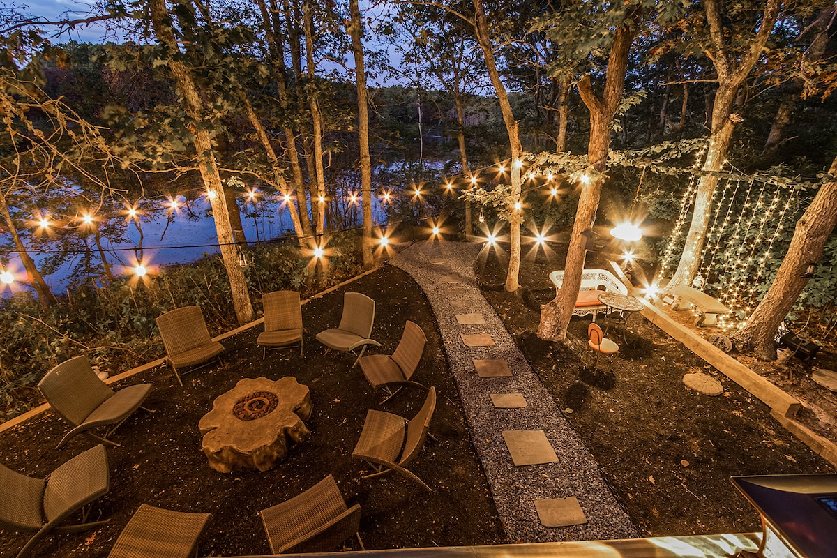 An enchanting outdoor area features a stone path leading to a fire pit surrounded by several chairs. String lights illuminate the scene, casting a warm glow on the natural landscape, with a serene pond view visible through the trees.