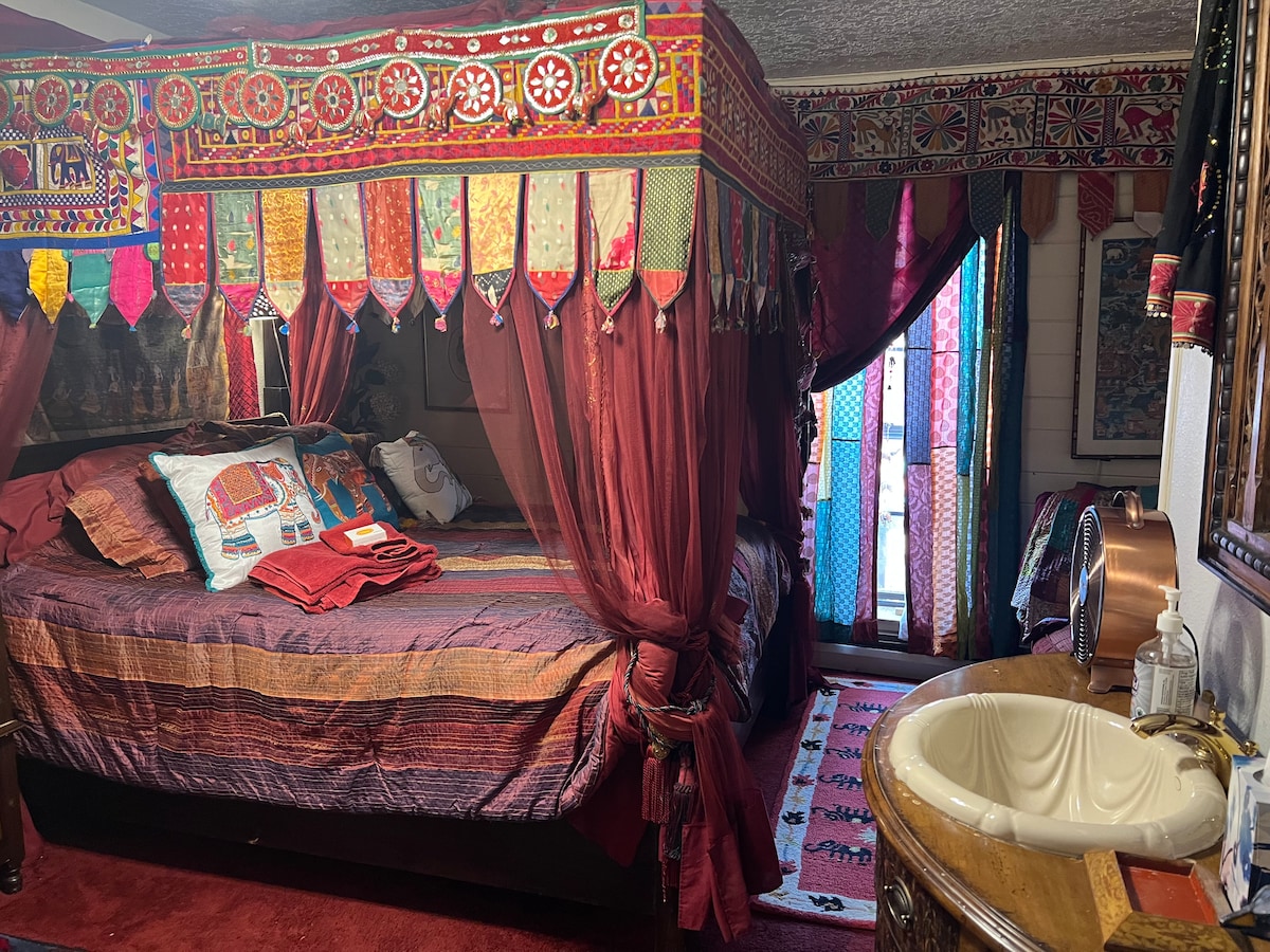 A vibrant canopy-style bed is surrounded by flowing drapes and colorful textiles. The room features bold patterns and decorative elements. A small vanity area is seen with a round sink, and light filters in through a beaded curtain, enhancing the inviting atmosphere.