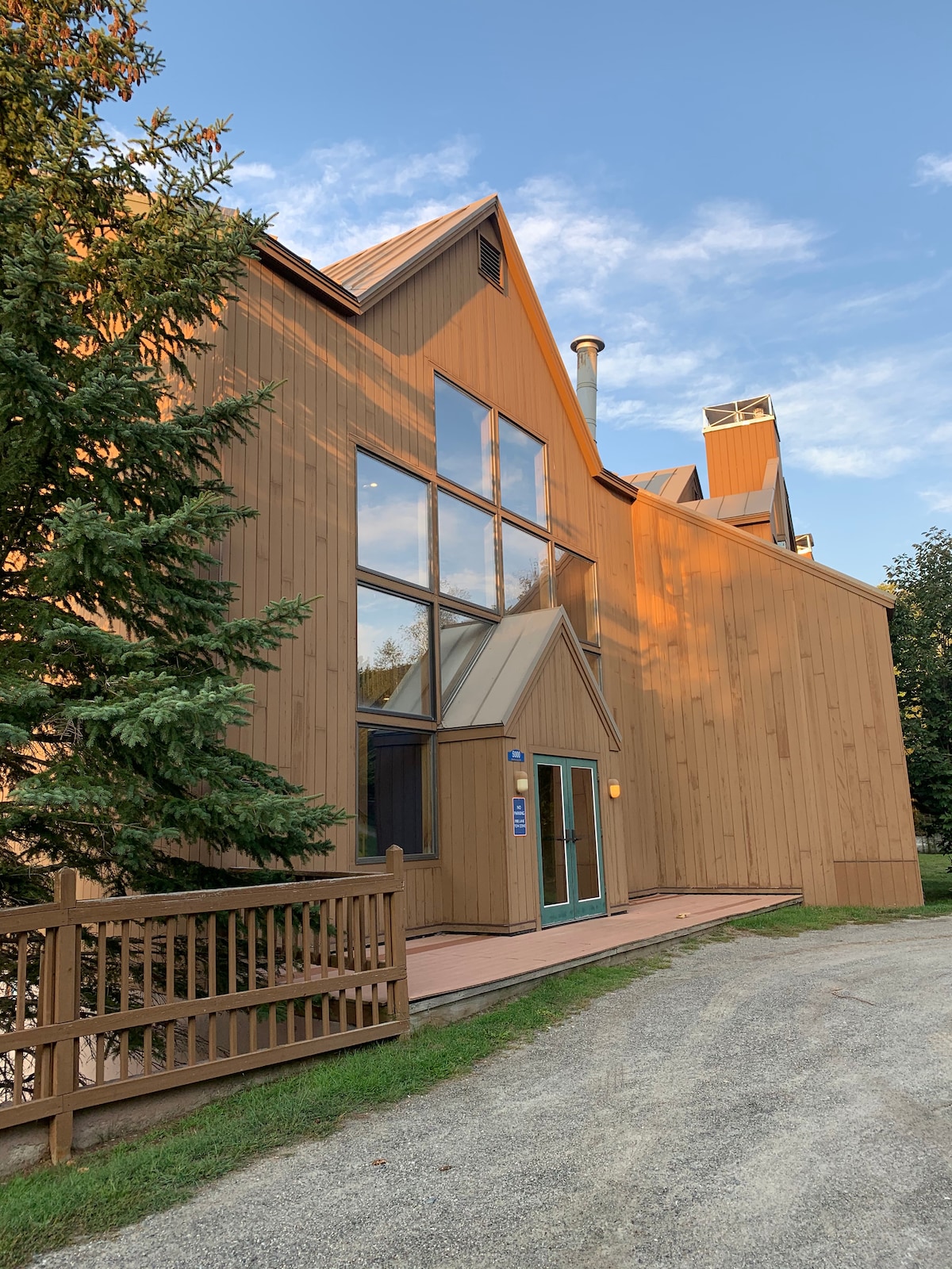 The exterior of the condo is showcased, featuring a combination of wood siding and large windows. Natural greenery surrounds the building, and a pathway leads to the entrance. A muted color palette reflects a harmonious blend with the inviting outdoor setting.