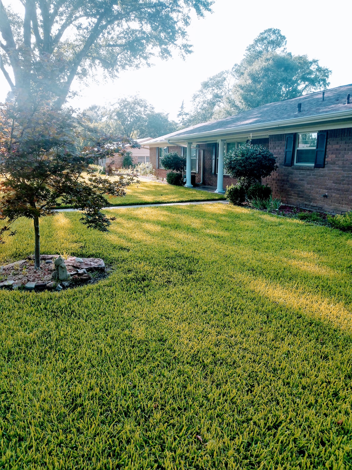 A well-maintained lawn features lush green grass, bordered by small bushes and trees. A tree stands prominently near a decorative stone circle, while the home's brick exterior is visible in the background, creating a welcoming and serene outdoor space.