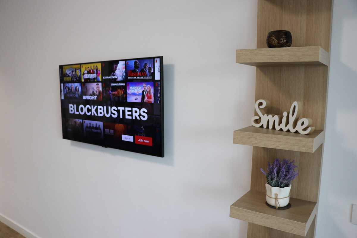 A flat-screen television displays a movie selection with the word 'BLOCKBUSTERS' prominently featured. Adjacent to it, a wooden shelf holds a small potted plant and a decorative item with the word 'Smile' inscribed. The wall is painted in a light hue, enhancing the modern aesthetic.