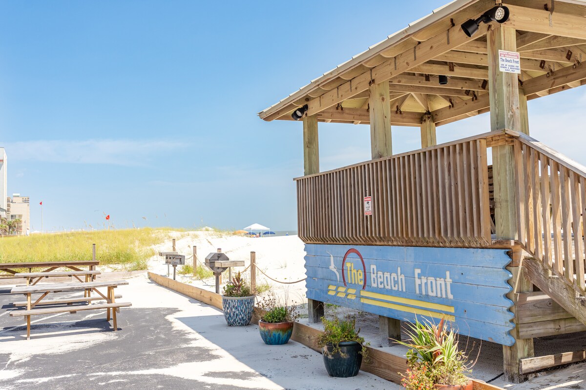 A wooden gazebo is featured prominently, painted in warm colors, with a sign indicating The Beach Front. Surrounding the gazebo, picnic tables are arranged on a sandy area adorned with potted plants. The beach and blue sky are visible in the background.