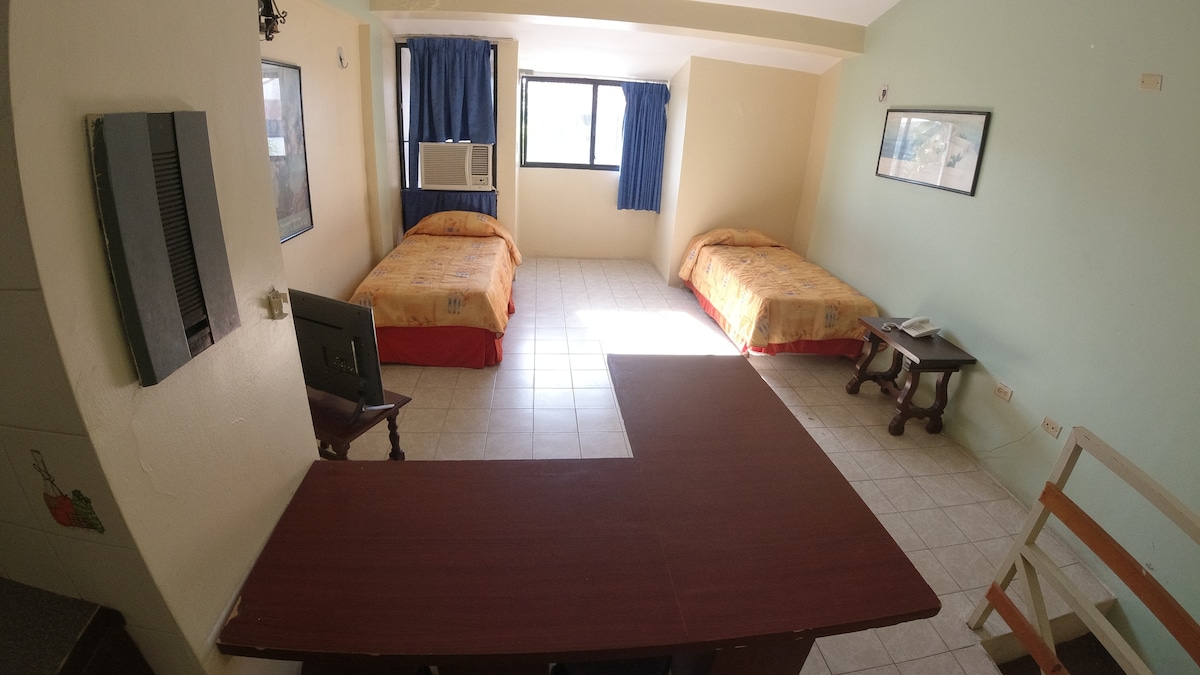 A bright and spacious room is furnished with two beds covered in light orange linens. A large window with blue curtains allows natural light to fill the space. A wooden desk with a chair is positioned near the entrance, and an air conditioning unit is mounted above.