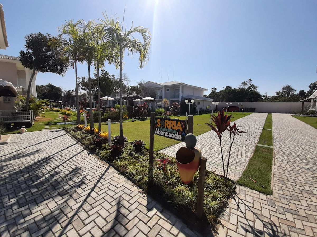 A well-maintained outdoor area is showcased, featuring a sign that reads 'Sorria' and decorative plants arranged along the pathway. Lush greenery surrounds the open space, with umbrellas providing shade over seating areas. Paved walkways lead to parking spaces, inviting a sense of community.