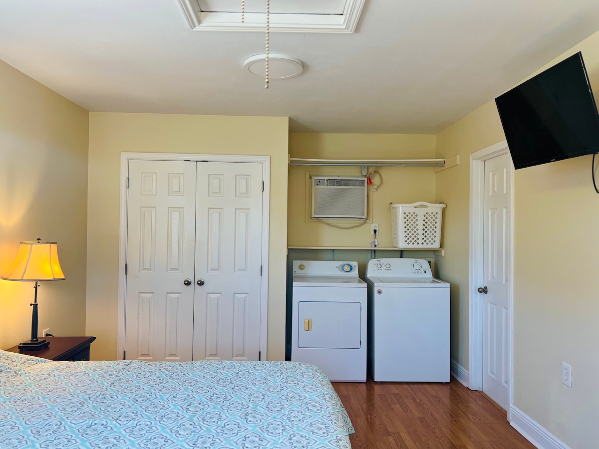 A cozy bedroom includes a spacious bed with a patterned comforter. A lamp provides soft light next to the bed. Laundry appliances are positioned against one wall, while a mounted television is fixed in the corner, ensuring convenient access and functionality.