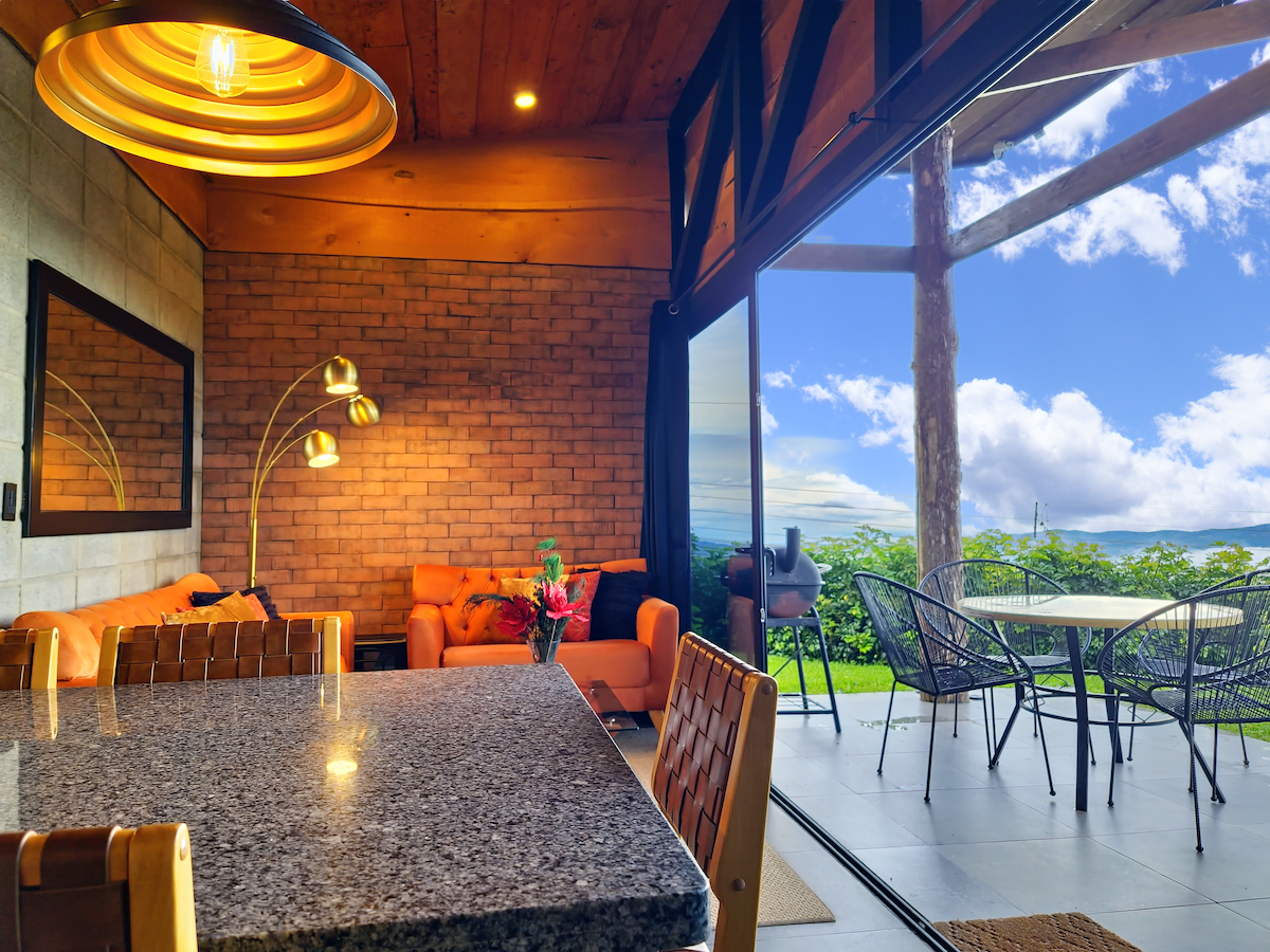 A cozy living space features a warm wood ceiling and a textured brick accent wall. A granite dining table occupies the foreground, while contemporary seating is arranged nearby. Large glass doors open to a patio with outdoor seating, providing scenic views of the surrounding landscape.